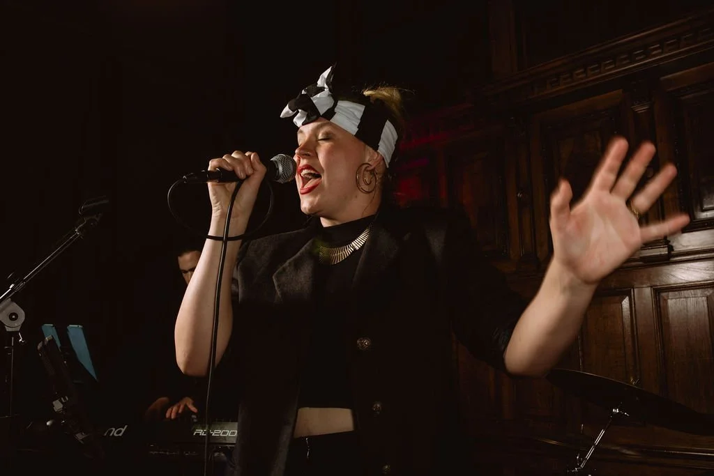 A woman singing into a microphone, wearing a black outfit and a headscarf, with a wooden background