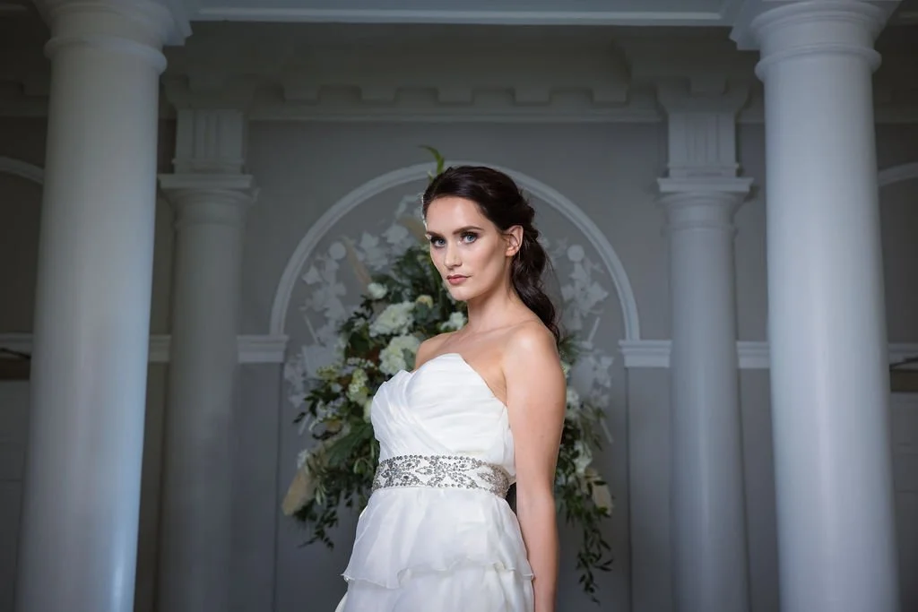 A woman in a white wedding dress standing in front of floral arrangements and white columns.