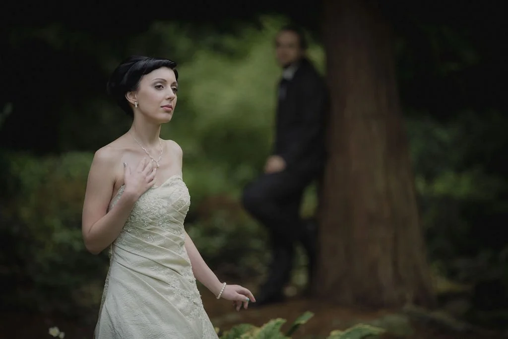 A woman in a strapless white dress stands outdoors with a serene expression, hand on her chest, in a wooded area, while a man in a dark suit leans against a tree in the blurred background.
