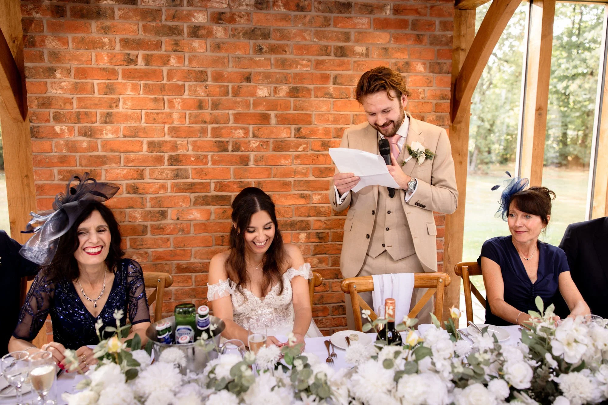 A man giving a speech at a wedding reception, standing behind a table with three women seated, all smiling, with a brick wall background and large windows showing green trees outside.