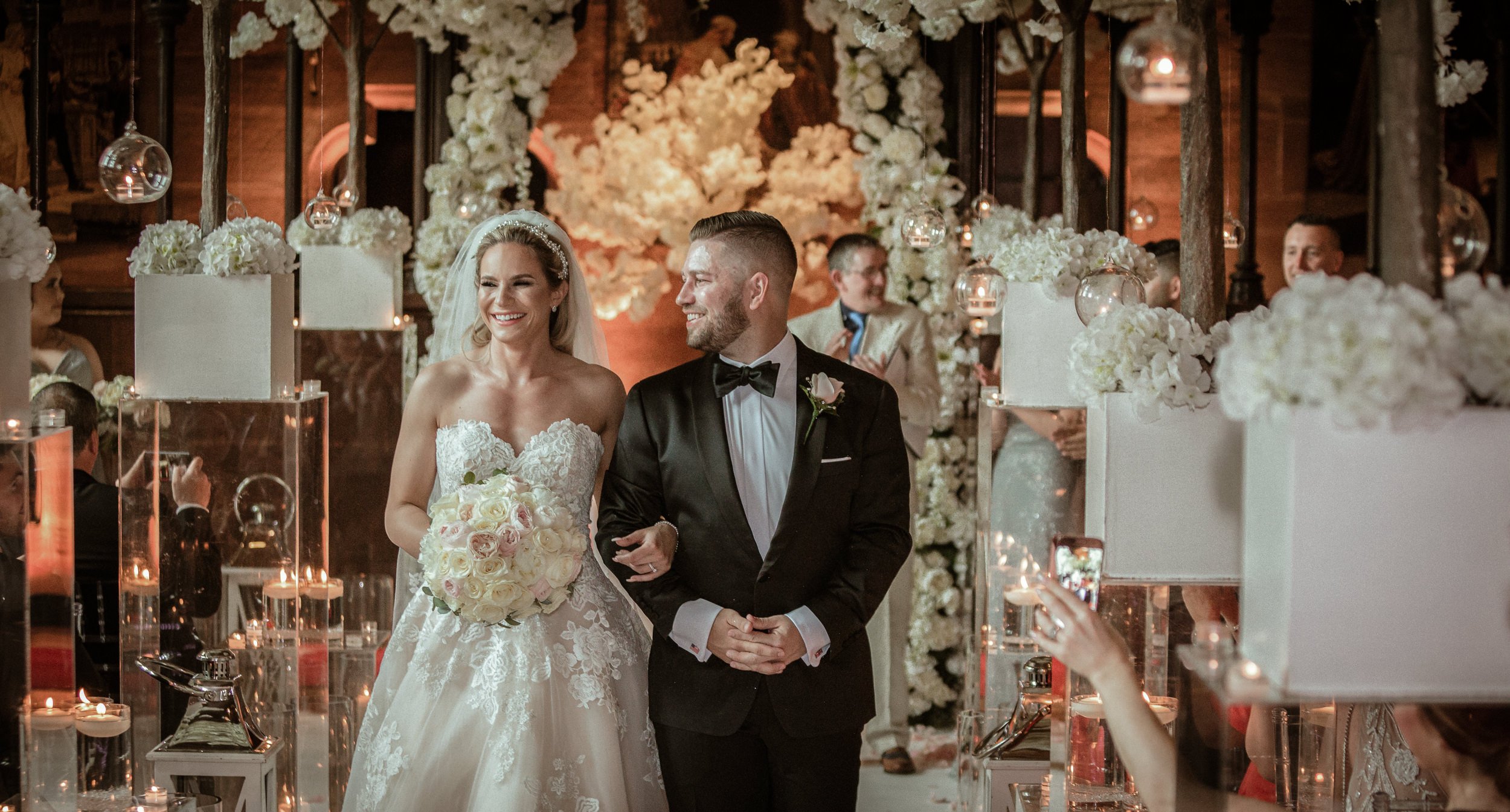 Bride and groom walking down the aisle at their wedding ceremony, surrounded by white flower arrangements and candles in glass holders in a warmly lit indoor venue.