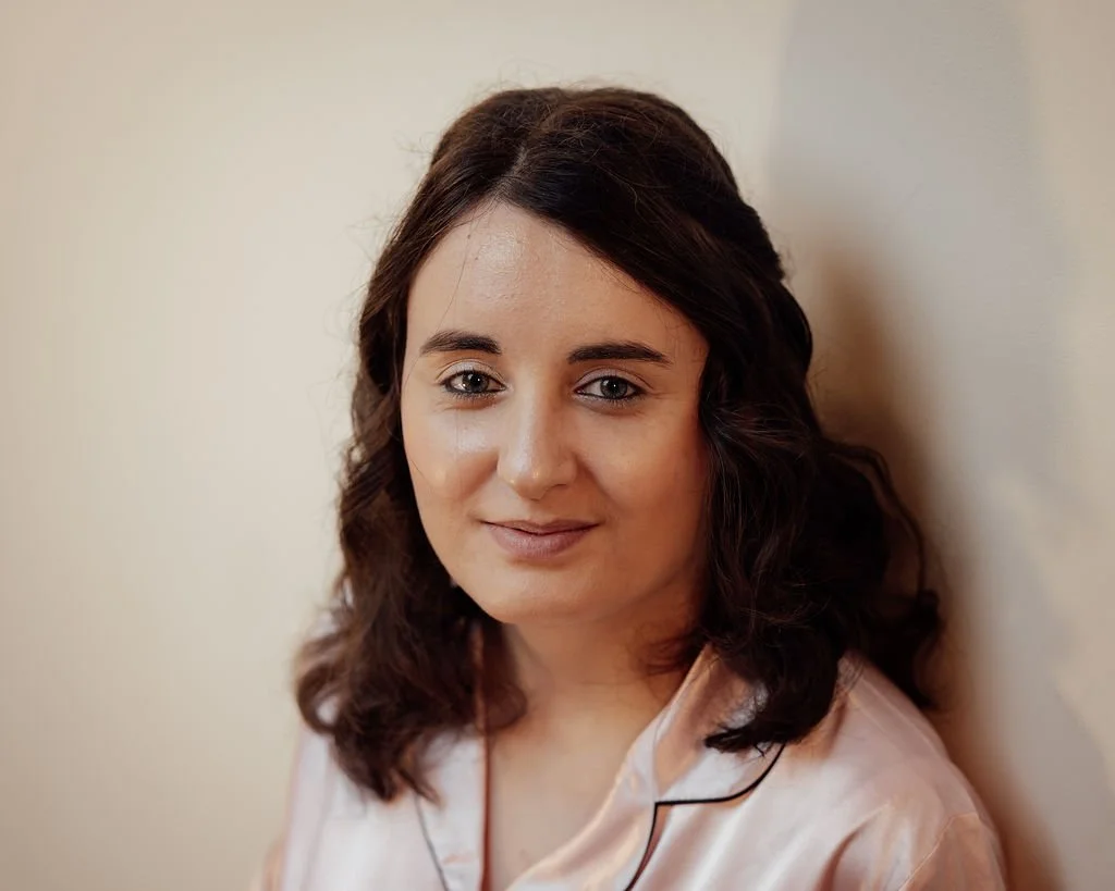A woman with shoulder-length dark brown hair and light skin smiling softly, wearing light pink satin pajamas, standing against a neutral-colored wall.