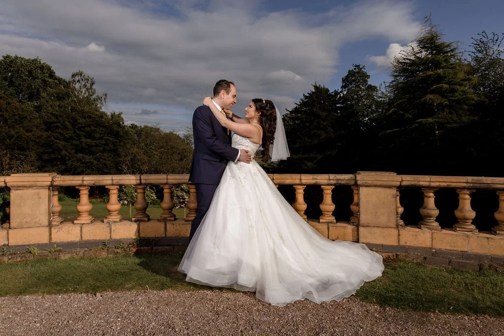 A bride and groom exchanging a kiss outdoors on a cloudy day, standing on a stone terrace with a scenic tree-filled background.