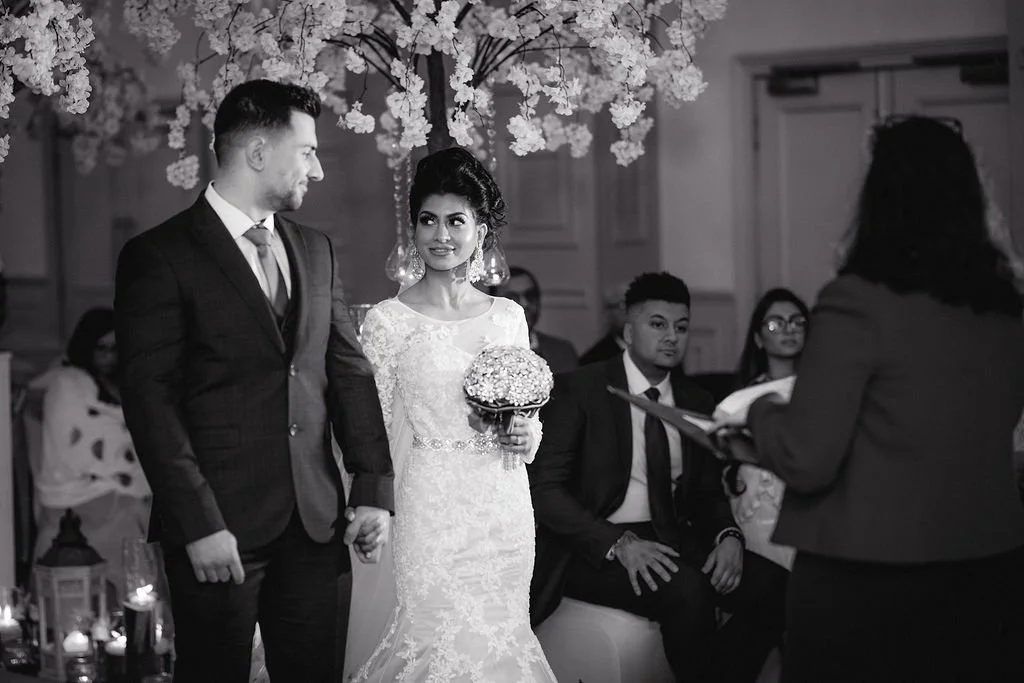 A bride and groom holding hands during their wedding ceremony, with the bride holding a bouquet, in a decorated venue with floral arrangements and seated guests.