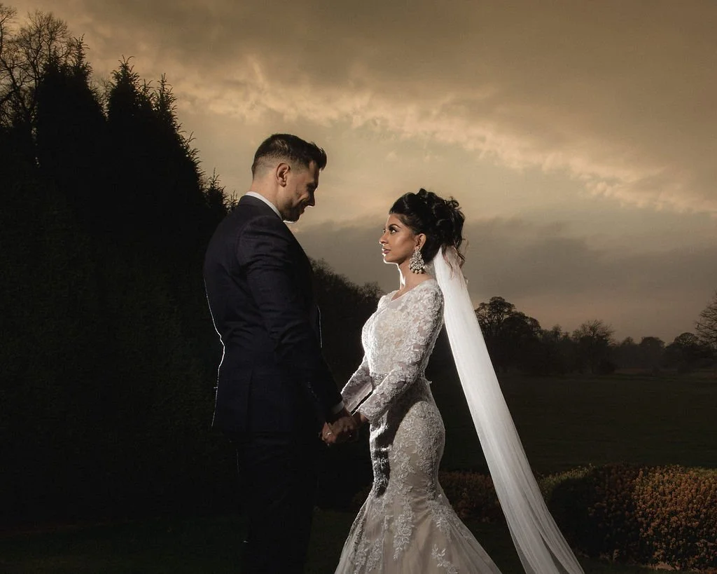 A bride and groom in wedding attire holding hands outdoors during sunset, with trees and a cloudy sky in the background.