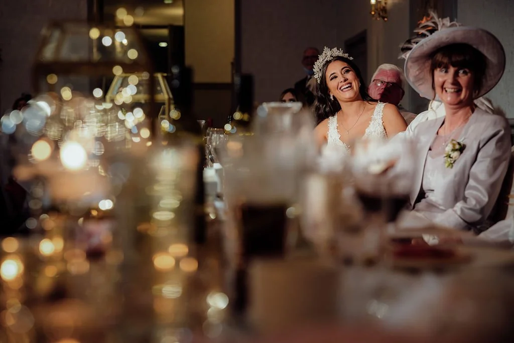 A bride in a white gown and floral crown laughing with an older woman in a large hat at a wedding reception.