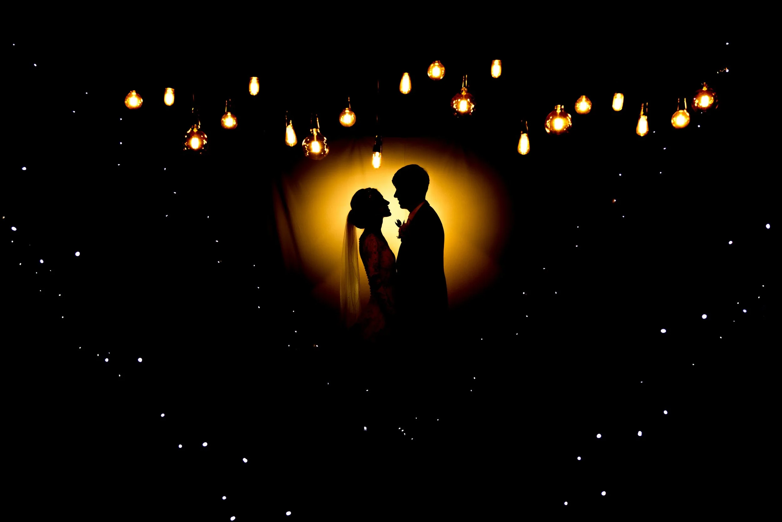 Silhouette of a bride and groom facing each other with a glowing yellow background, surrounded by hanging vintage Edison bulbs and small white star-like lights.