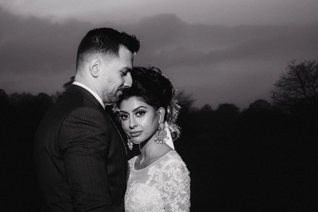 A couple dressed in formal wear, standing closely together outdoors during dusk, with a dark, cloudy sky and leafless trees in the background.