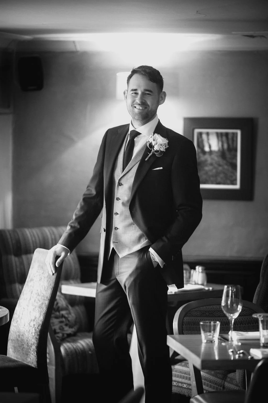A man dressed in a tuxedo, smiling, standing indoors with a boutonniere on his lapel, near a dining table with glassware and a chair.