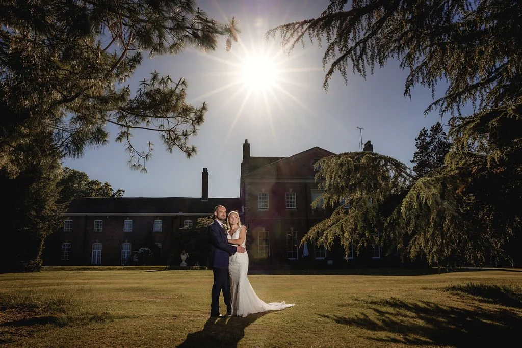 A newlywed couple standing on a grassy lawn in front of a large, historic brick mansion, with the sun shining brightly overhead and surrounded by tall trees.