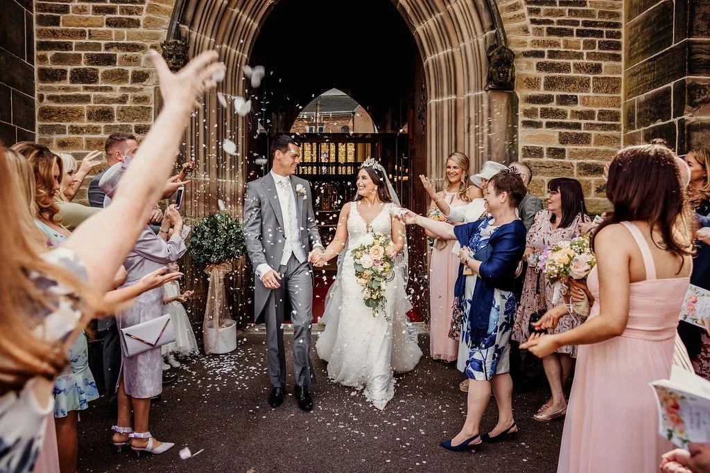 Bride and groom walking through a crowd of wedding guests throwing flower petals outside a church entrance.
