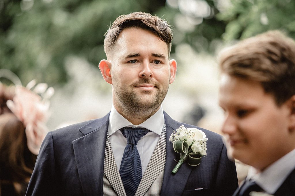A man with short brown hair and a beard, wearing a dark suit, a white shirt, and a patterned tie, at a wedding with a boutonniere of white flowers and green leaves on his lapel, standing outdoors with blurred greenery in the background.