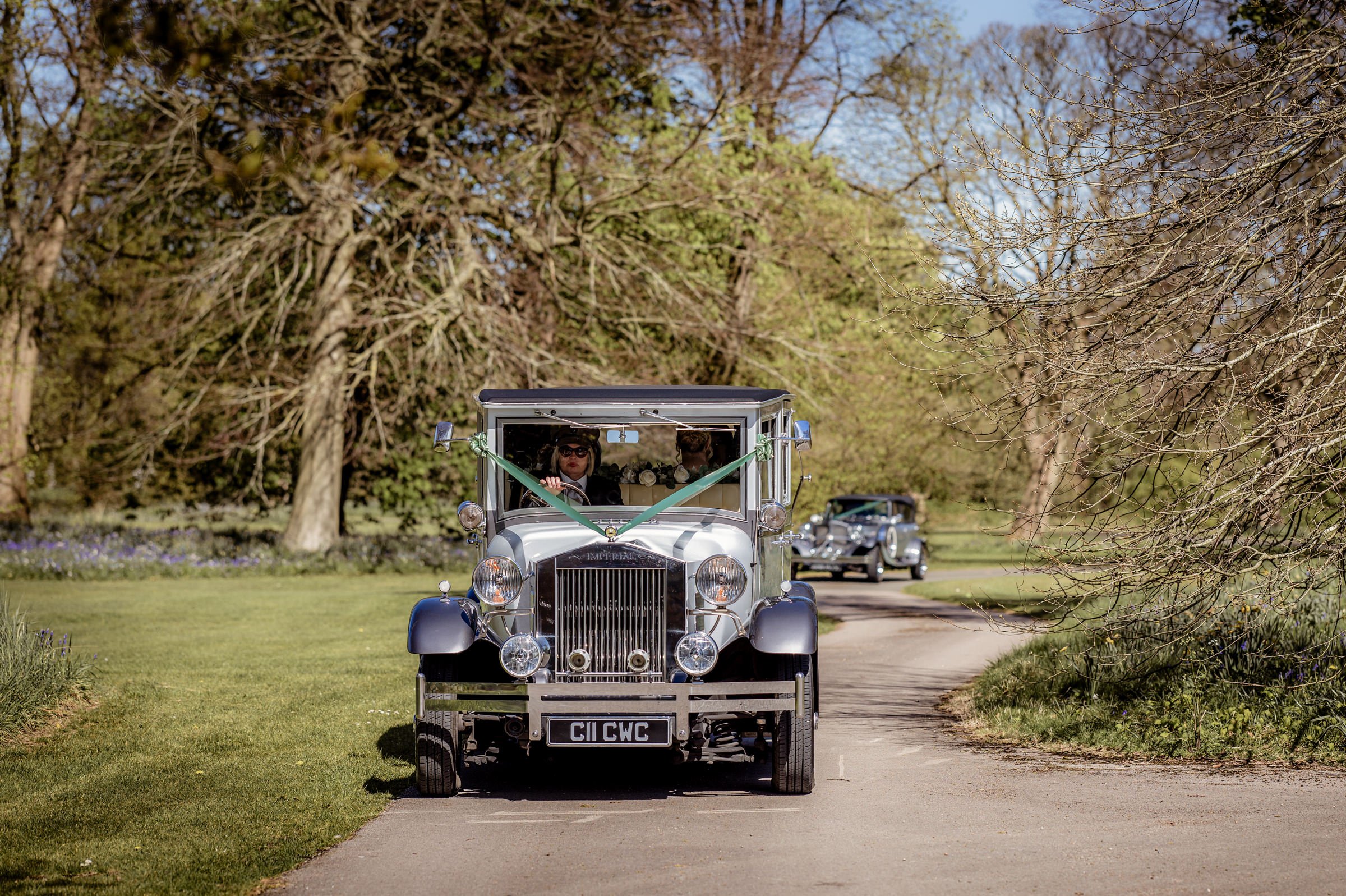 Vintage white car driving on a country road with trees and another similar car in the background.