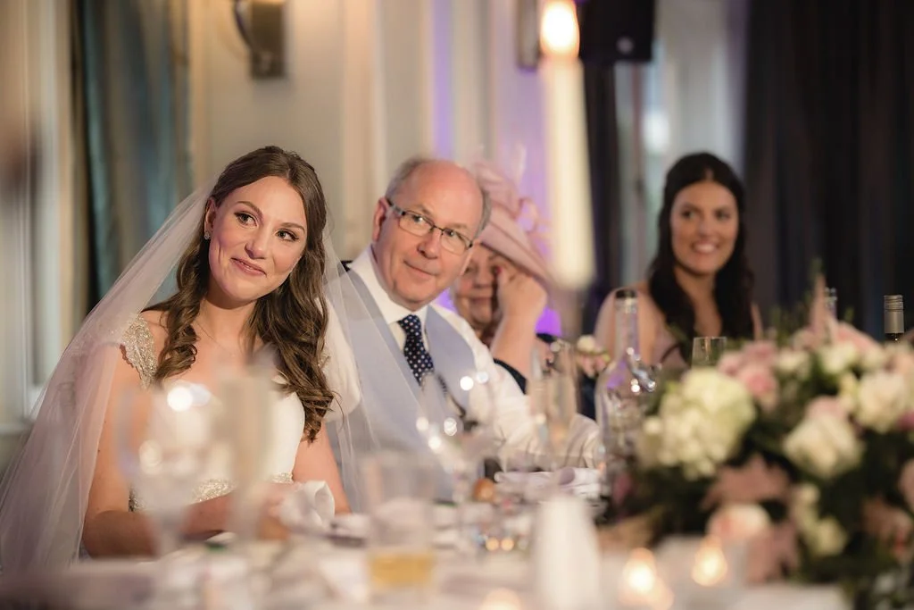 Bride with long brown hair in wedding dress, and older man with glasses and a polka dot tie sitting at a table during a wedding reception, with a woman in black dress smiling in the background, and floral arrangements on the table.