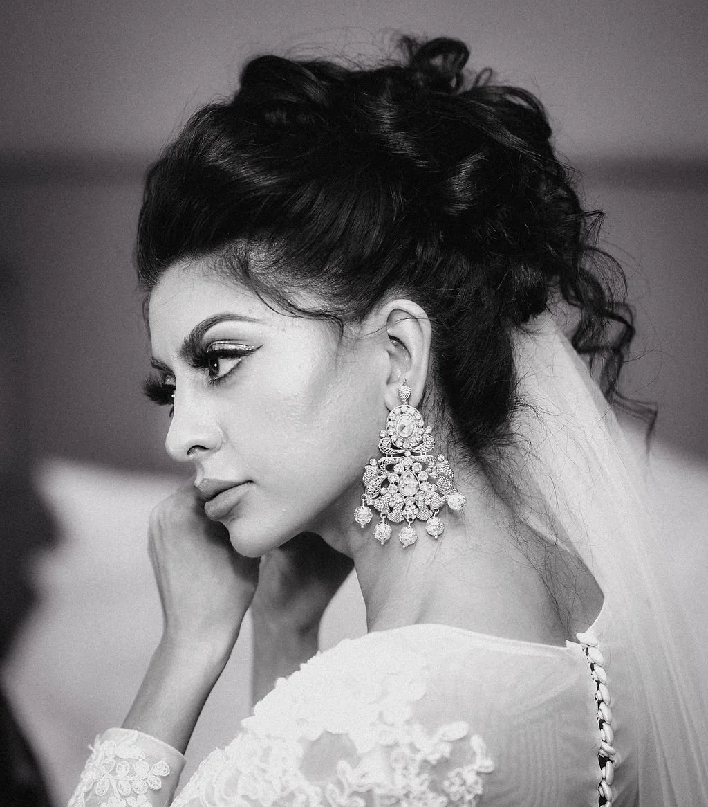 A black-and-white photo of a woman with elaborate earrings, styled hair, and makeup, looking to the side with her hand near her face.