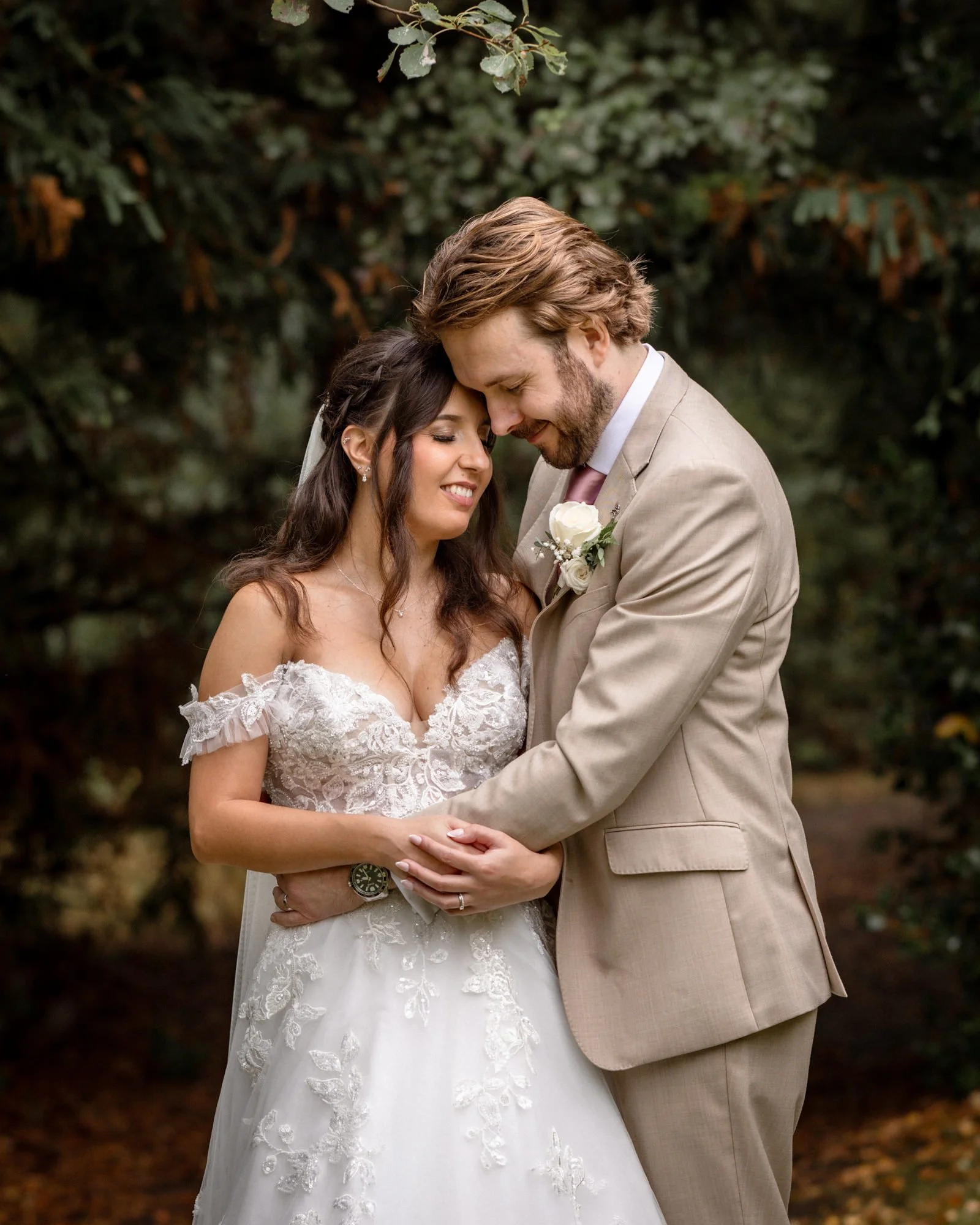 A bride and groom embrace outdoors, with their foreheads touching and eyes closed, dressed in wedding attire, standing among trees with dark green foliage.