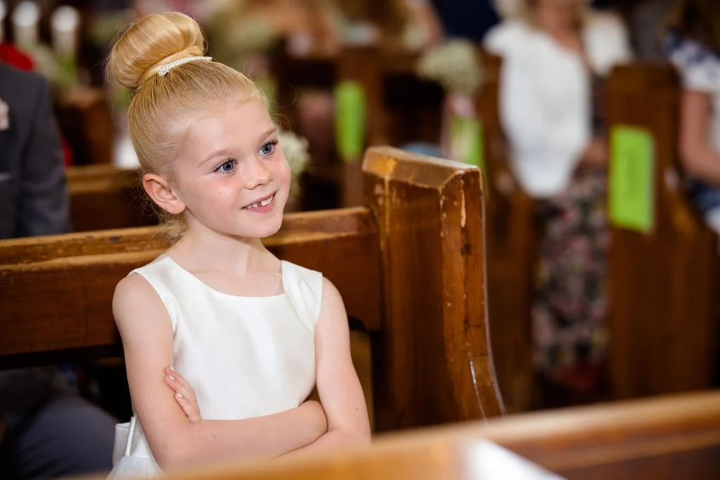A young girl with blonde hair in a bun sitting with crossed arms in a church, smiling and looking ahead.
