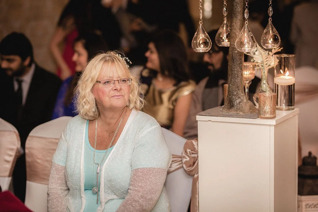 An older woman with blonde hair wearing glasses, a light-colored cardigan, and jewelry, sitting at a formal event with dim lighting, surrounded by other guests in elegant attire, with decorative lighting and candles in the background.