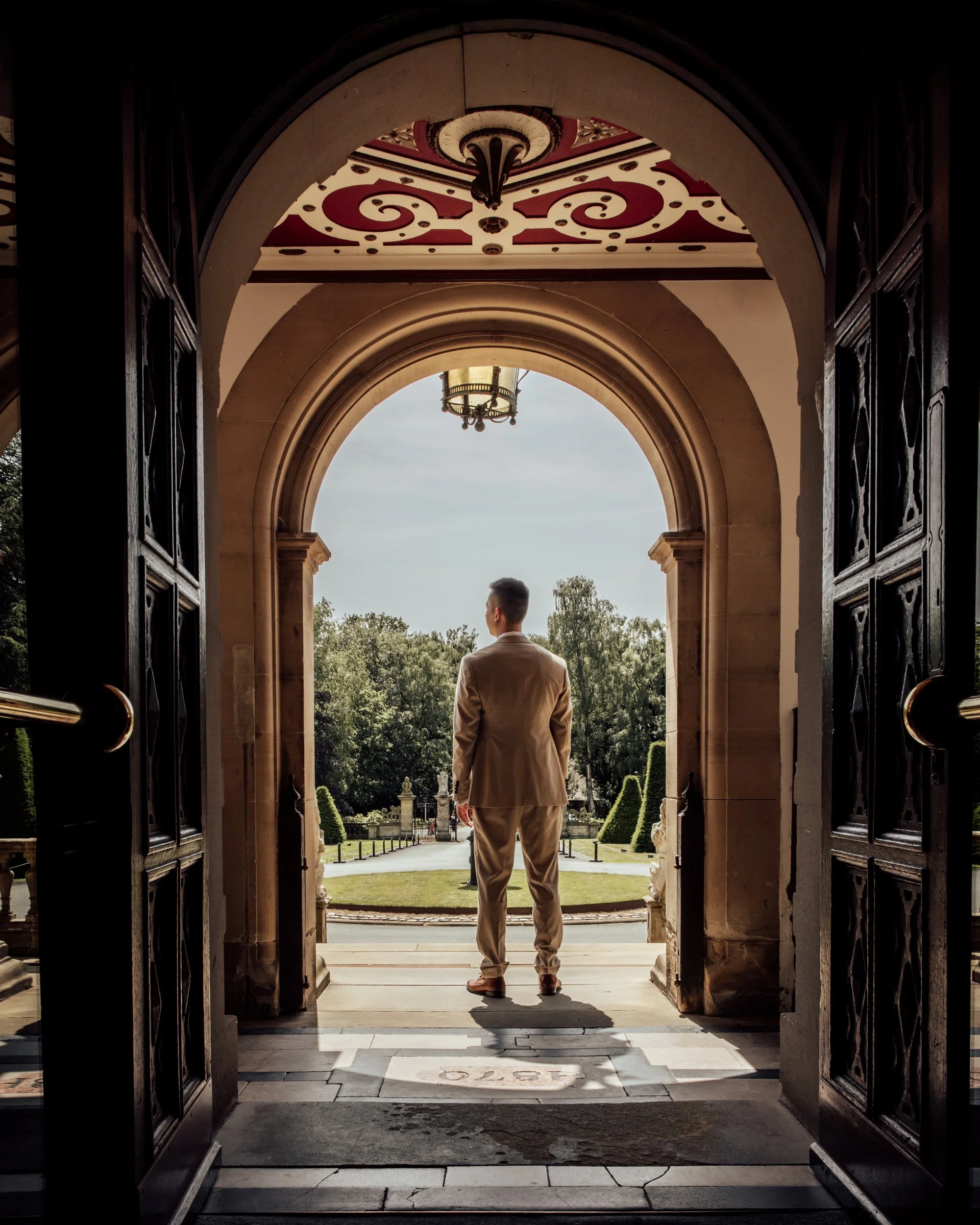 A man in a beige suit standing in the doorway of a building, looking out towards a garden with trees and a fountain, with a view of the sky and a chandelier hanging from the ceiling.