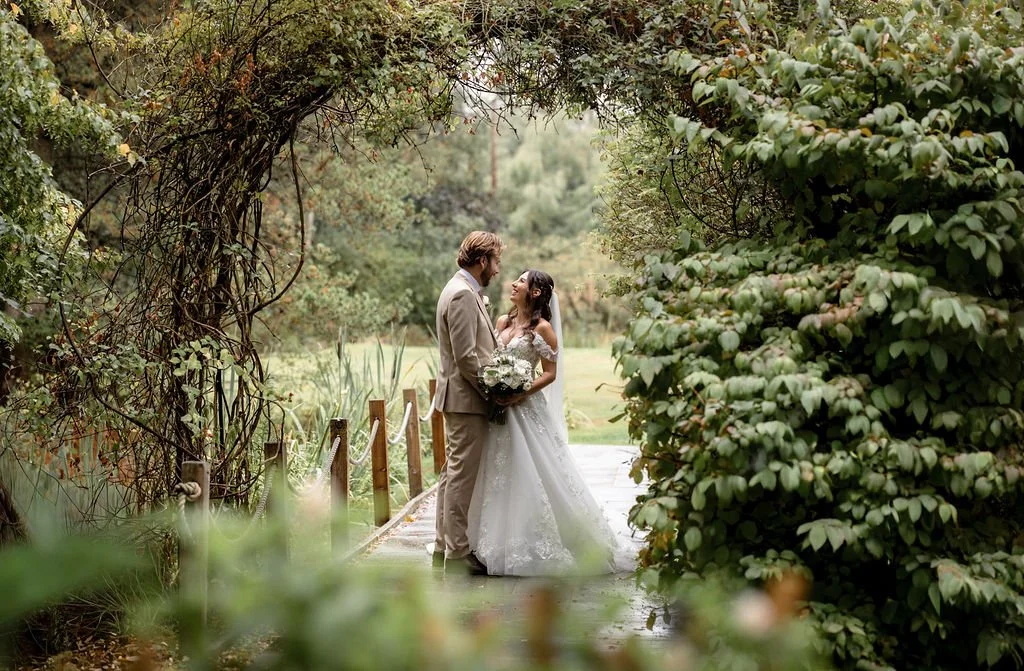 A bride and groom standing on a small wooden bridge surrounded by lush green foliage.