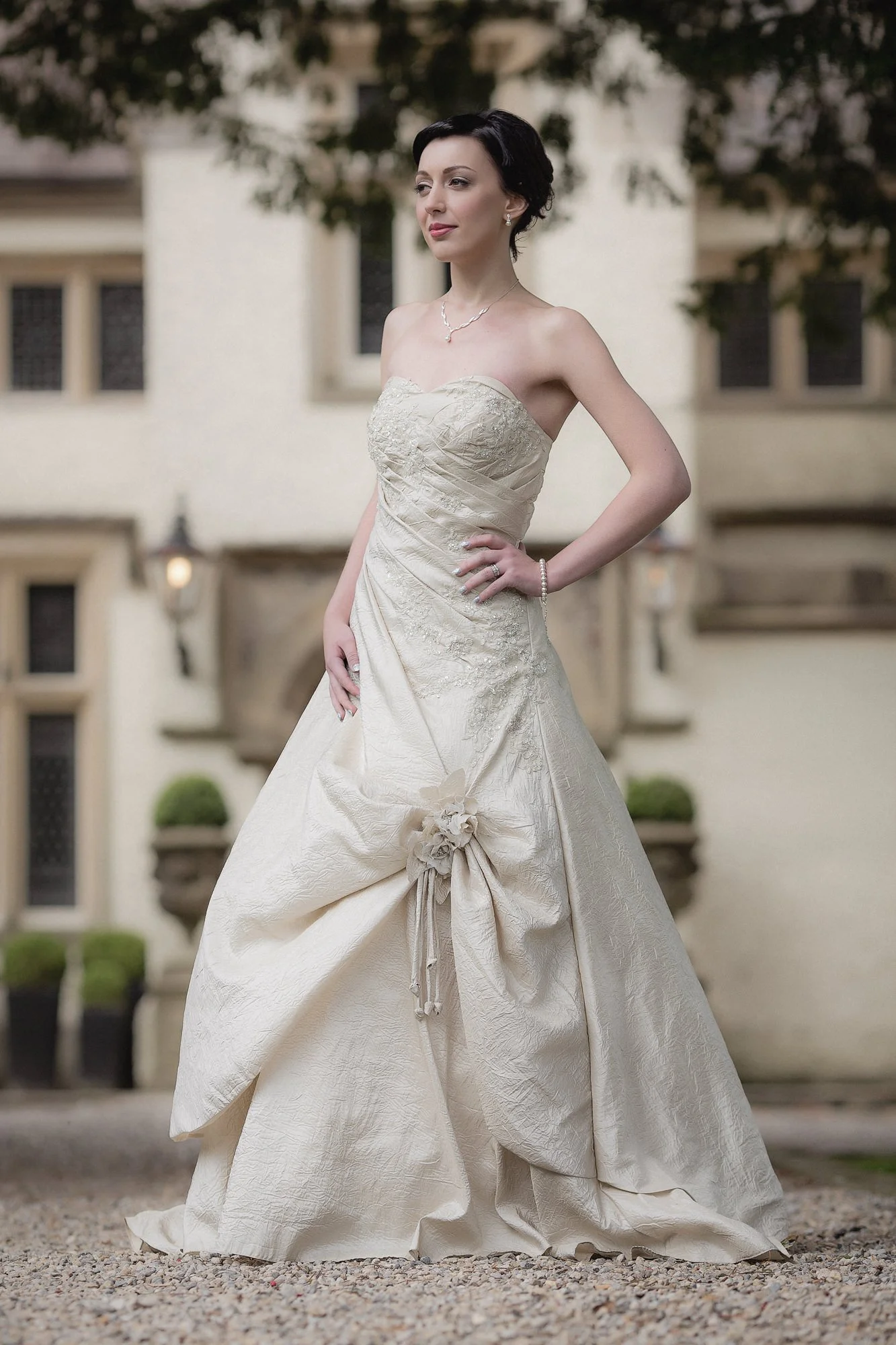 A woman in a strapless wedding dress standing outdoors in front of a building with windows and floral planters.