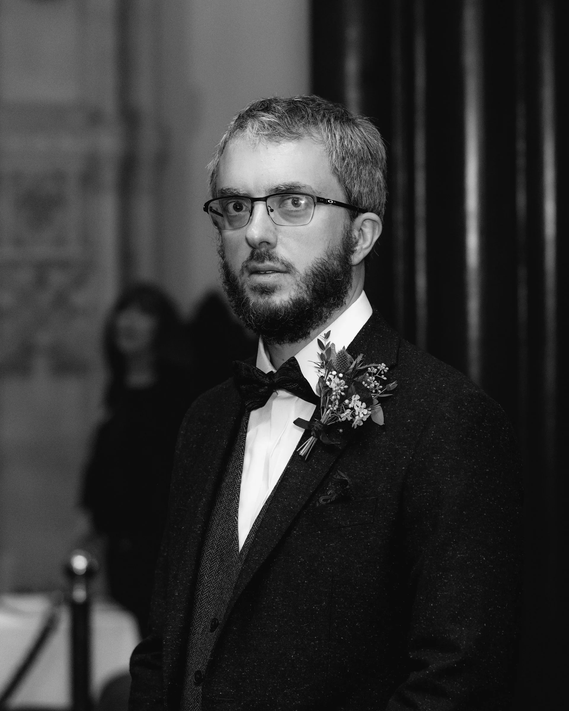 A man with glasses and a beard wearing a tuxedo with a boutonnière at a formal event.