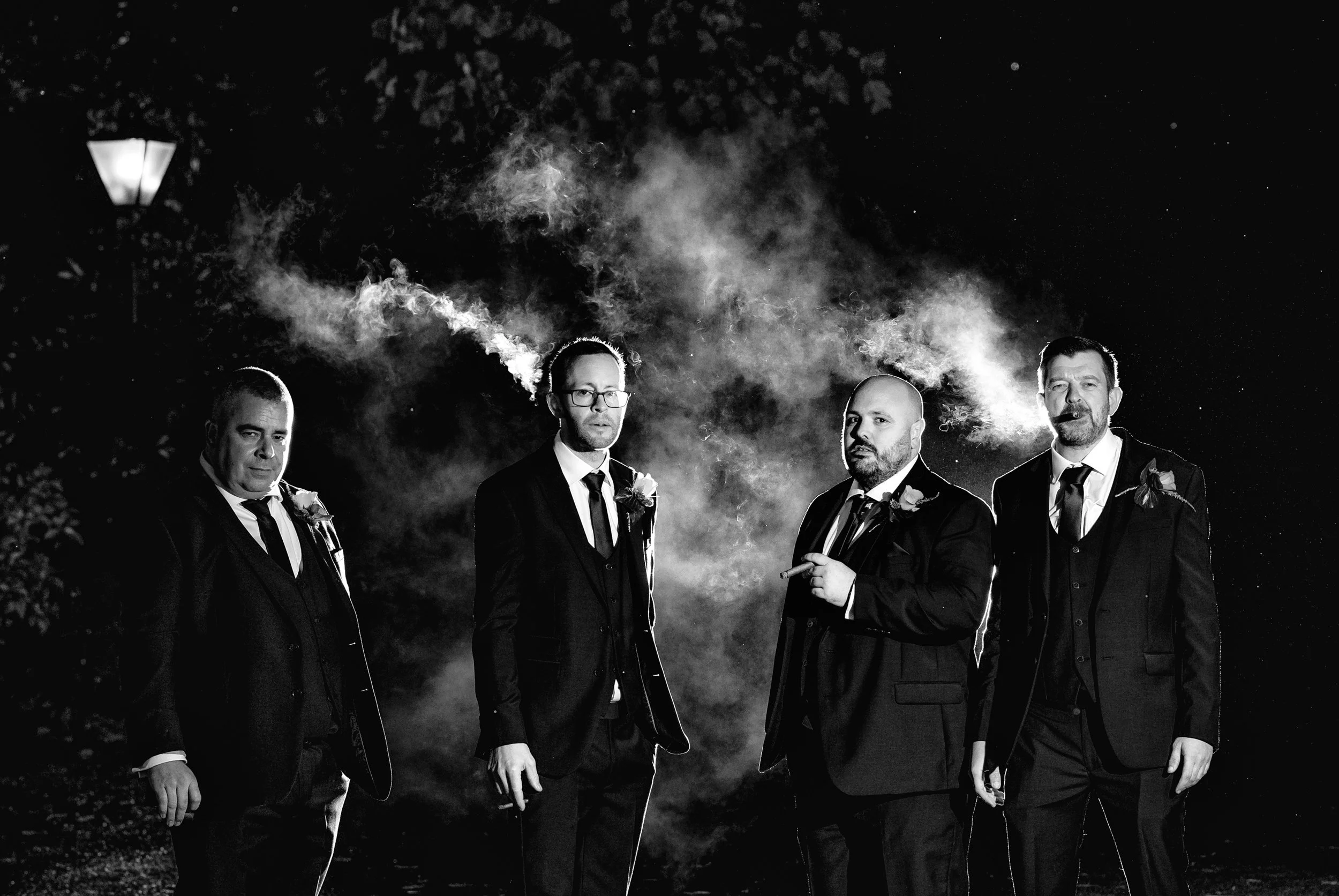 Black and white photo of four men in suits, standing outdoors at night, with street lamps in the background. Two of the men are smoking cigars, and smoke is visible around their heads.