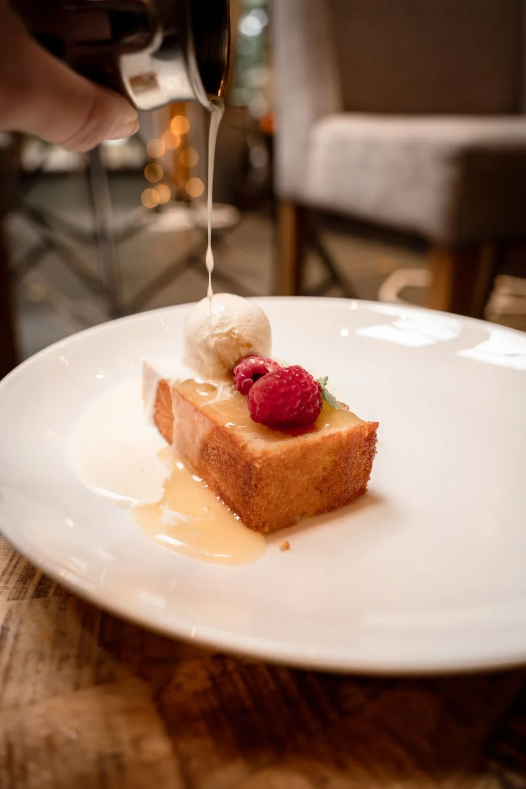 A slice of cake topped with raspberries and a scoop of ice cream, with caramel sauce being poured over it.