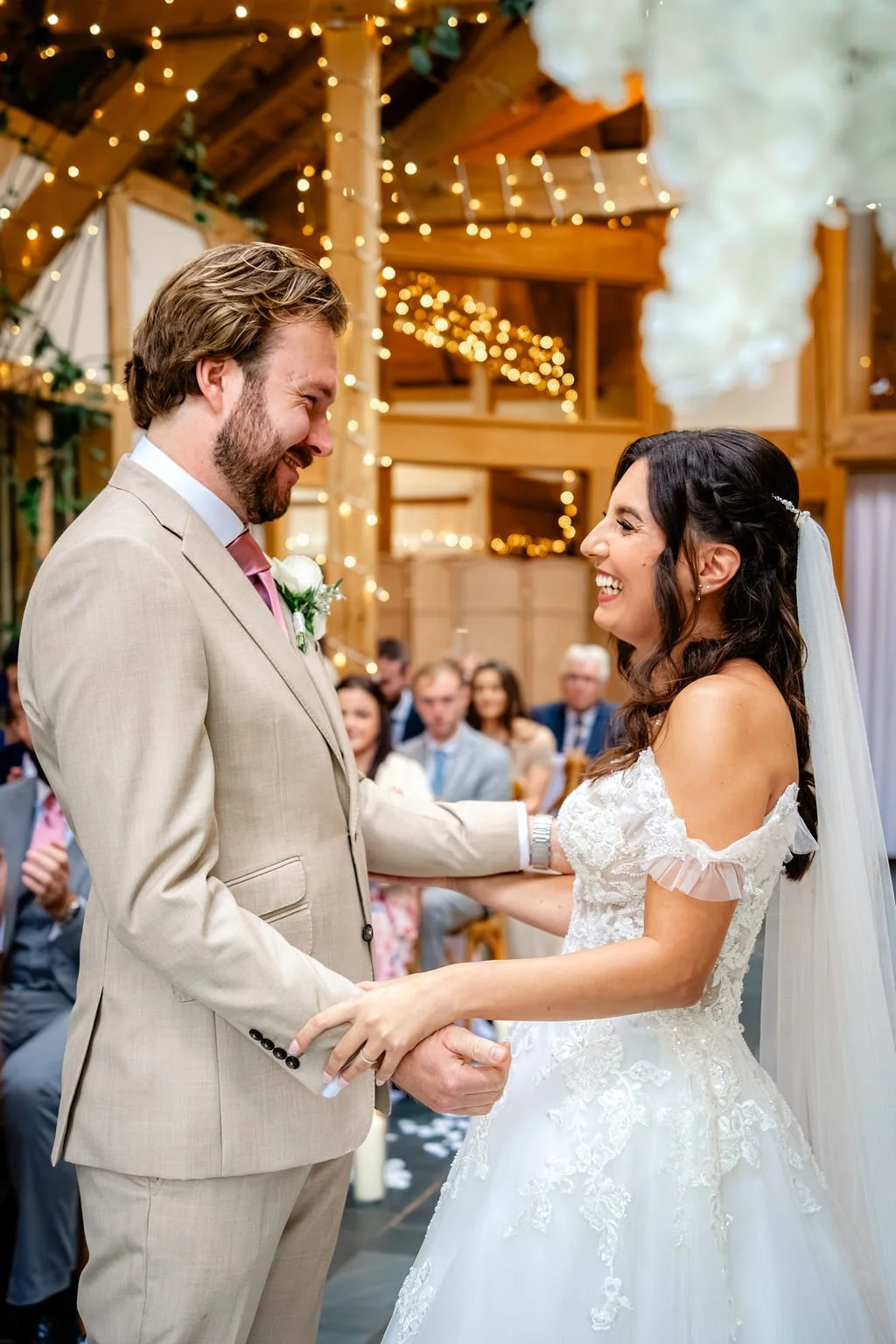 A bride and groom smiling at each other during their wedding ceremony inside a decorated wooden venue with string lights.