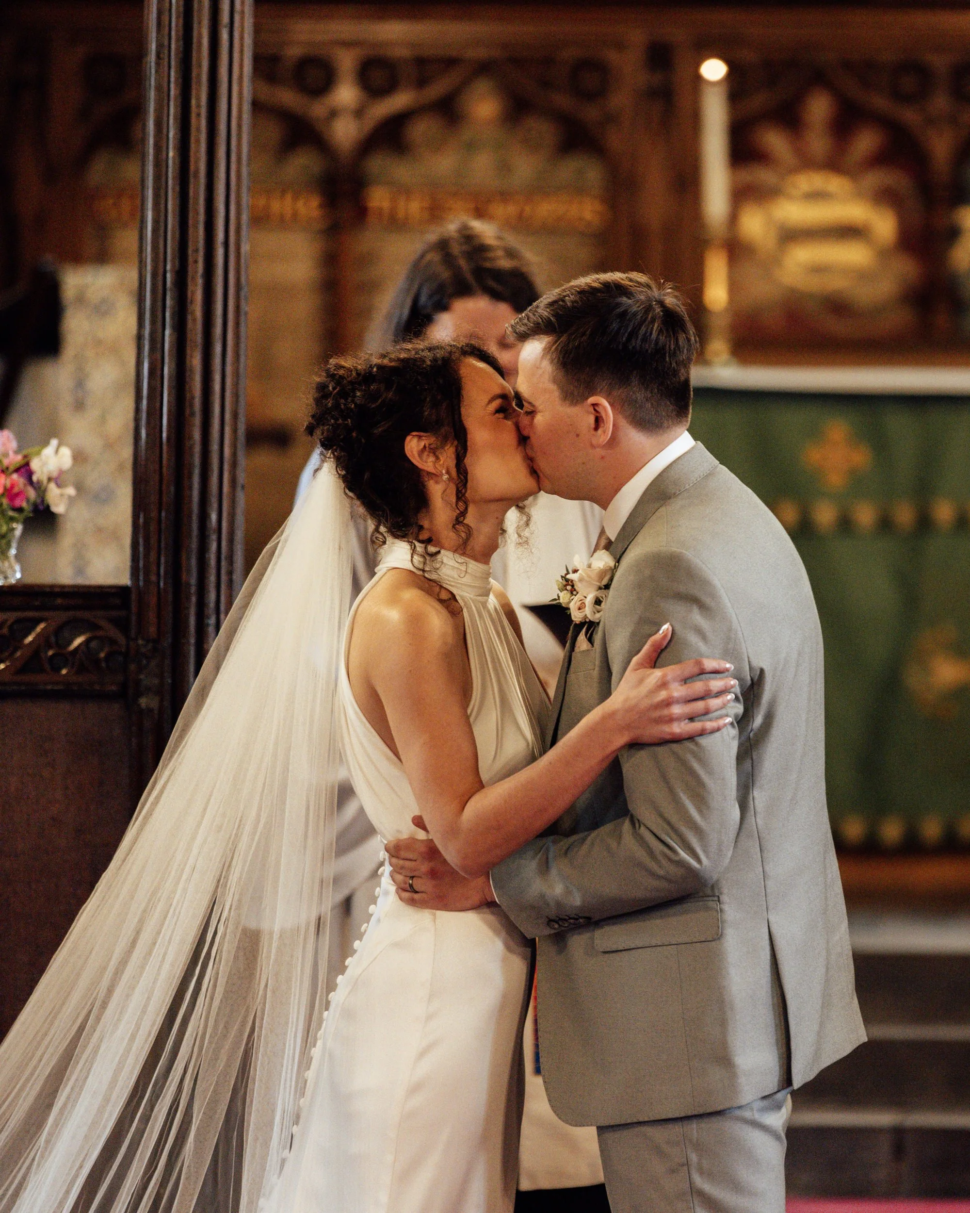 A bride and groom share a kiss during their wedding ceremony inside a church, with a woman officiant in the background.