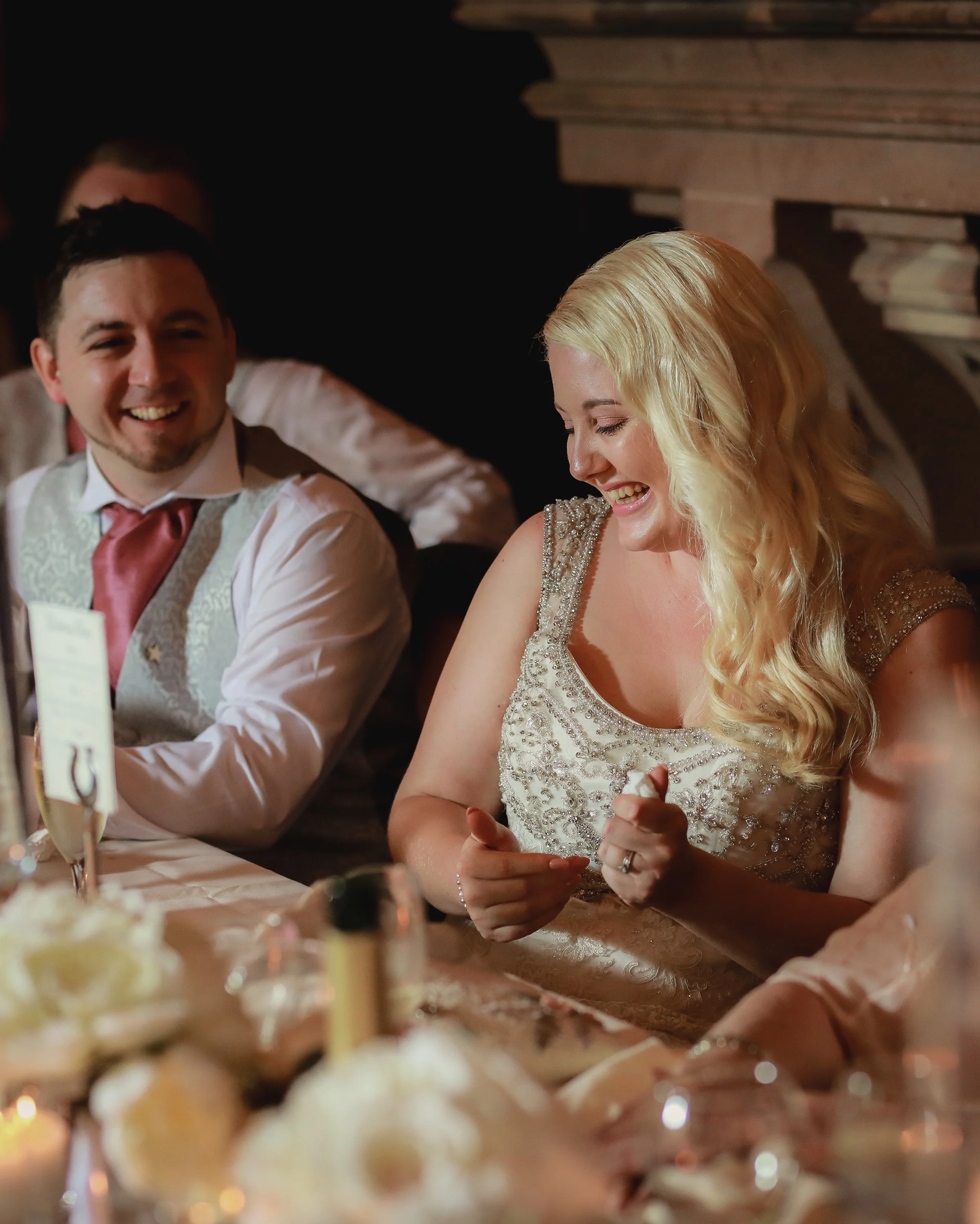 A bride with long blonde hair laughing and sitting at a wedding reception table, wearing a beaded wedding dress, alongside a smiling groom in a vest and tie, with dinnerware and floral decorations on the table.