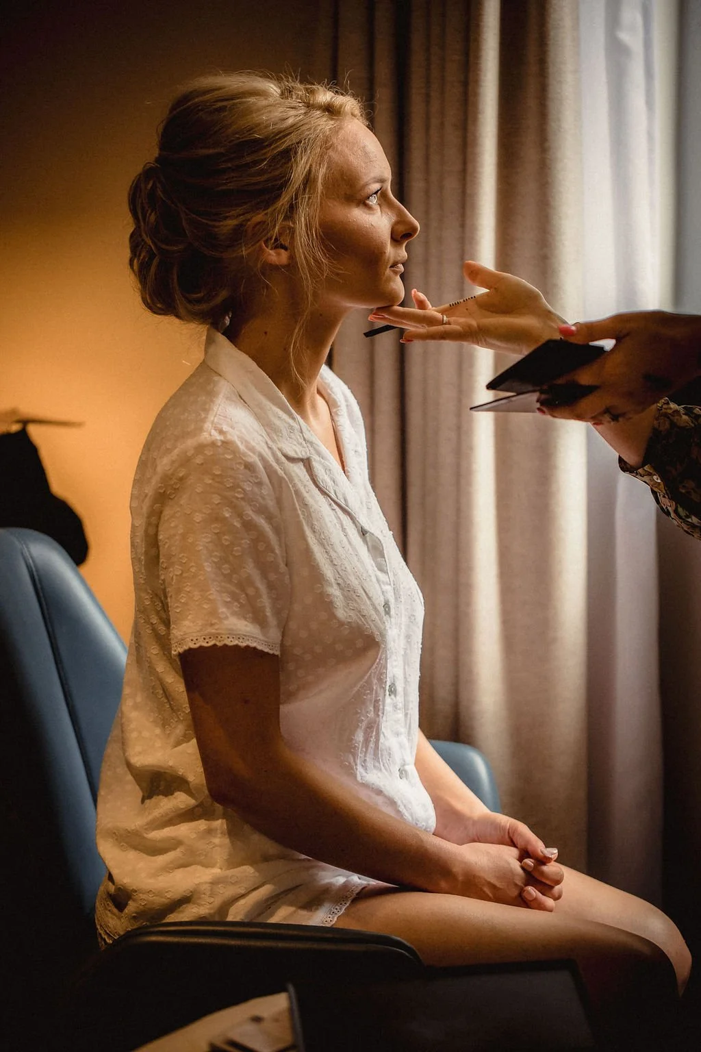 A woman with styled blonde hair is sitting on a chair wearing a white lace top. A person with manicured nails and dark clothing is applying makeup or touching her face, holding a makeup brush and a small mirror. The woman is looking straight ahead wi