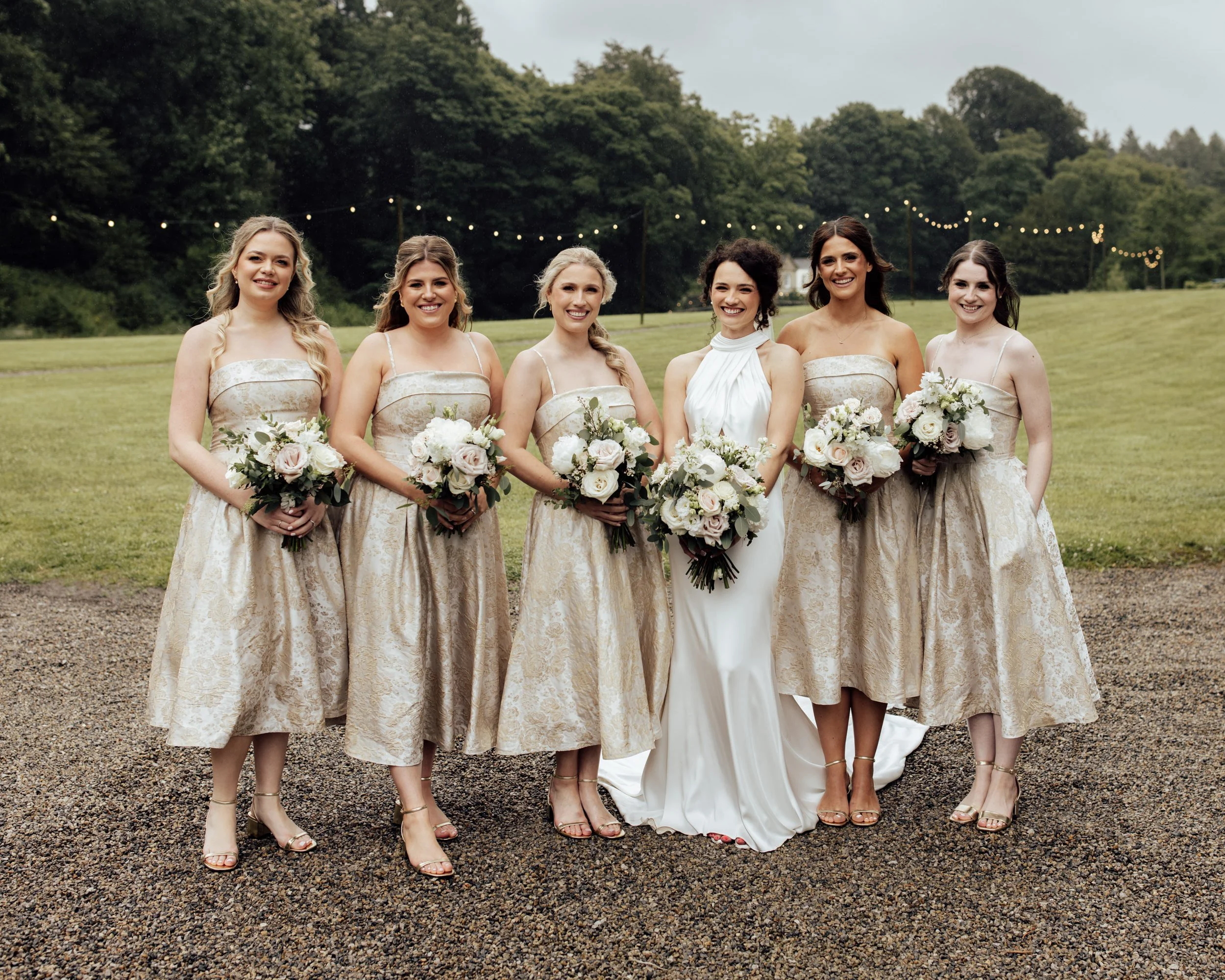 Six women dressed in matching champagne-colored dresses holding bouquets, standing outdoors on a grassy field with trees and string lights in the background, smiling at the camera.