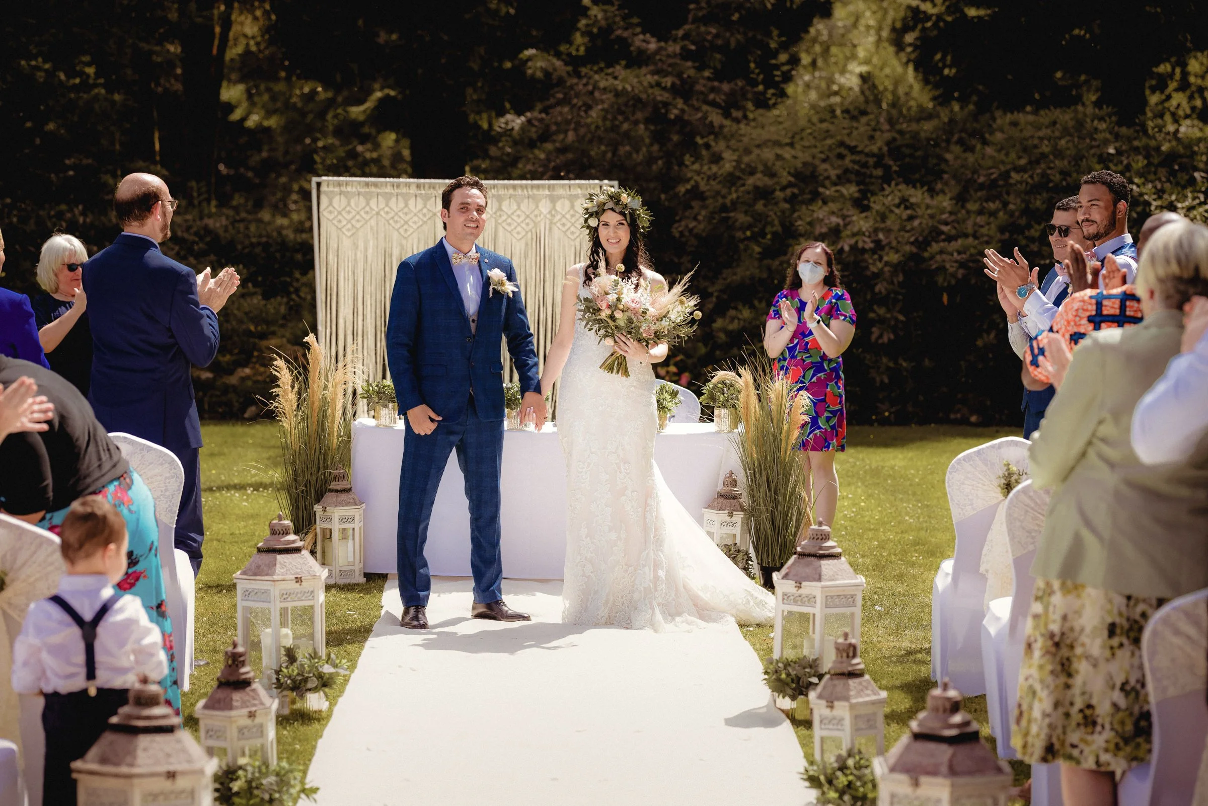 A bride and groom standing hand-in-hand at their outdoor wedding ceremony, surrounded by applauding guests on a sunny day.