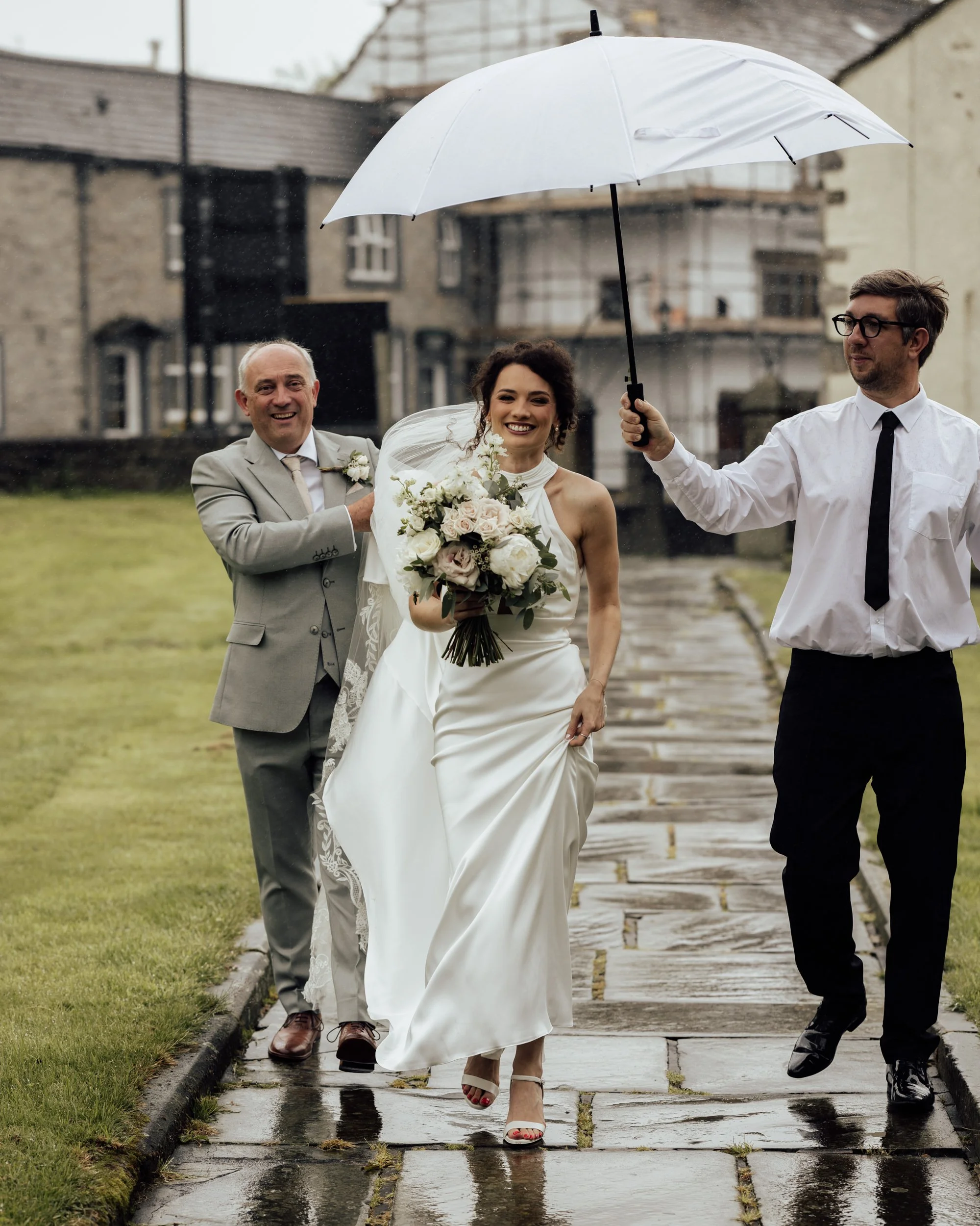 A bride in a white wedding dress holding a bouquet of flowers walking on a wet stone pathway while a man holds an umbrella over her, with another man walking behind her on a rainy day in an outdoor setting with old buildings in the background.