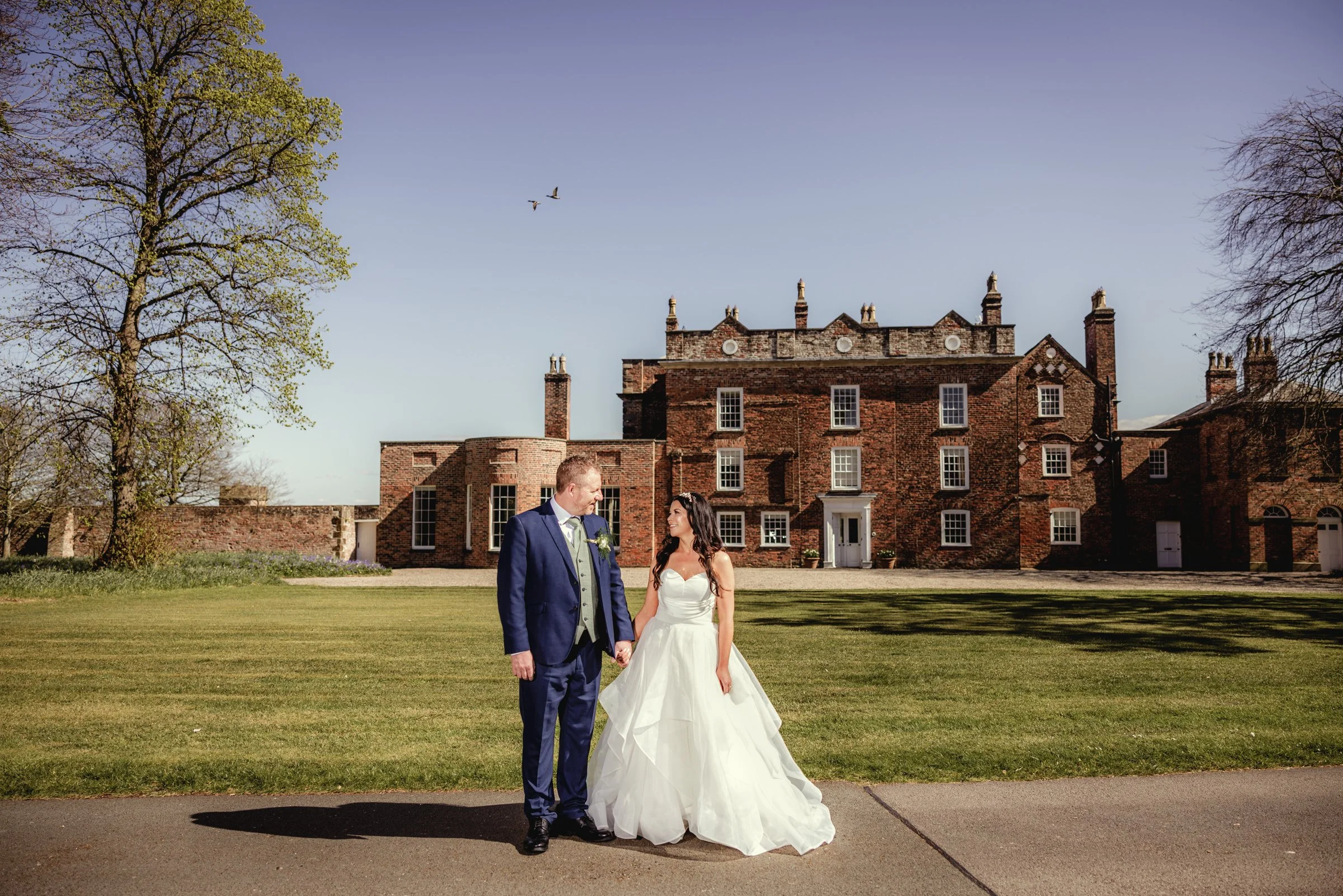 A bride and groom stand hand in hand in front of a large brick mansion under a clear sky. The bride is wearing a white wedding gown, and the groom is dressed in a dark blue suit. There are trees and a green lawn surrounding them.