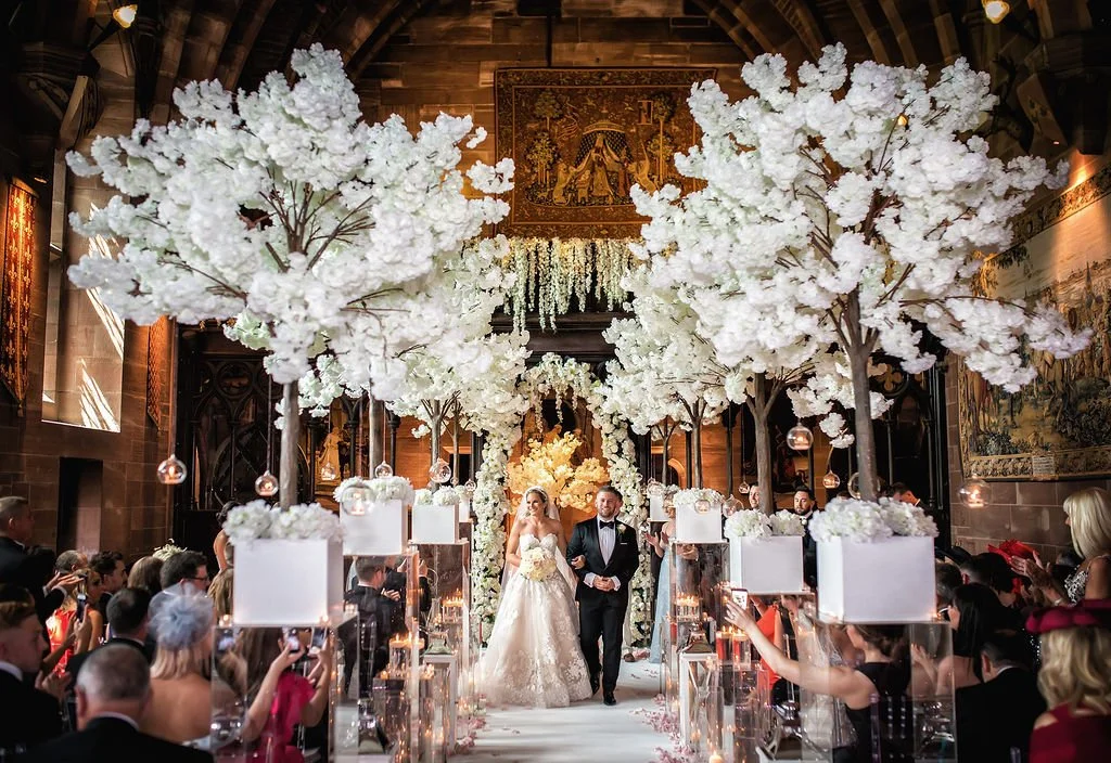 Bride and groom walking down the aisle in a beautifully decorated church with white floral trees and candles, surrounded by wedding guests.