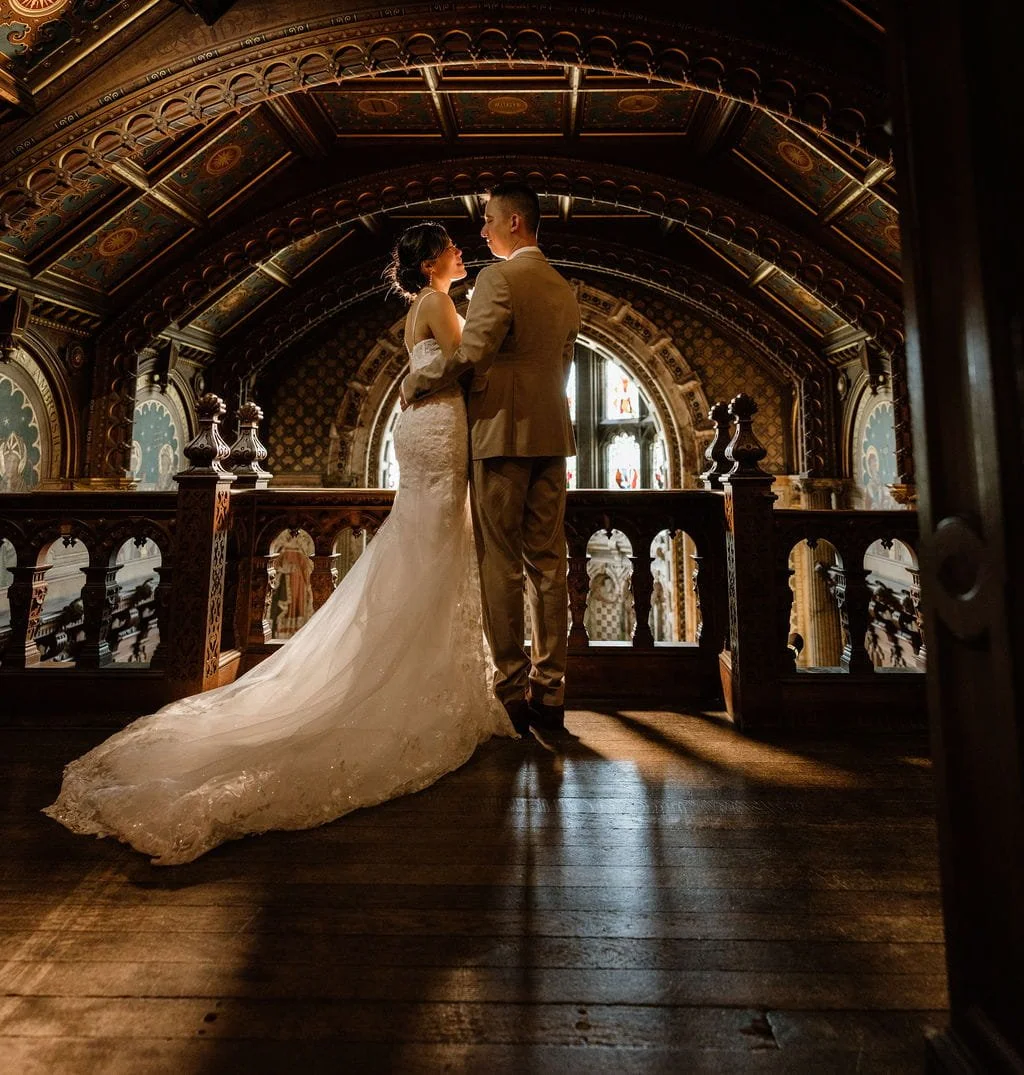 A bride and groom standing close together in a historic building with stained glass windows and ornate woodwork, illuminated by warm light.