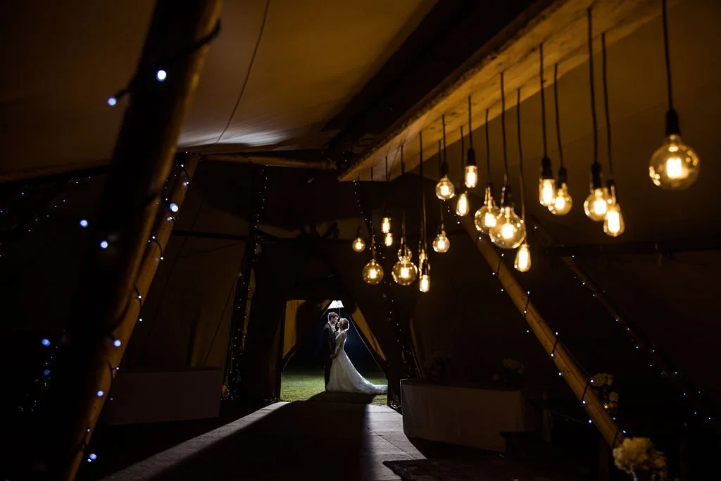 A bride and groom standing together inside a dimly lit tent illuminated by hanging Edison bulbs, with decorative string lights, viewing each other at the entrance.