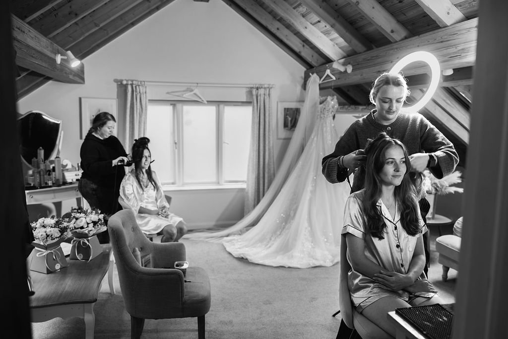 A woman is getting her hair styled by a hairstylist in a room with a sloped ceiling, with a wedding dress hanging in the background. Another woman is sitting on a chair, and a makeup artist is working on her. The room contains a vanity with makeup an