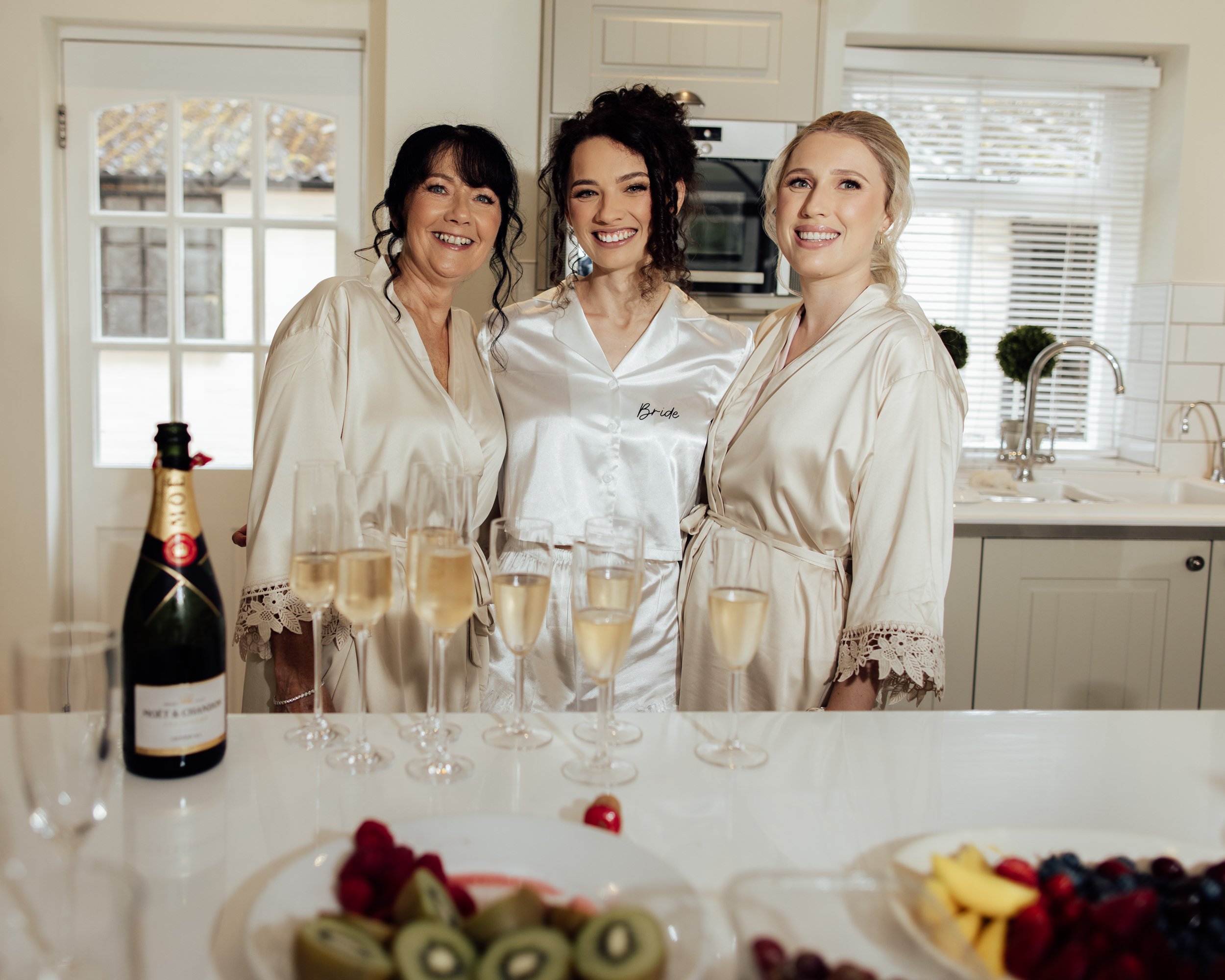 Three women smiling, dressed in satin robes, standing in a kitchen with a window and wooden cabinets. One woman has a 'Bride' embroidered robe, suggesting a wedding celebration. In the foreground, there are glasses of champagne and a plate of assorte