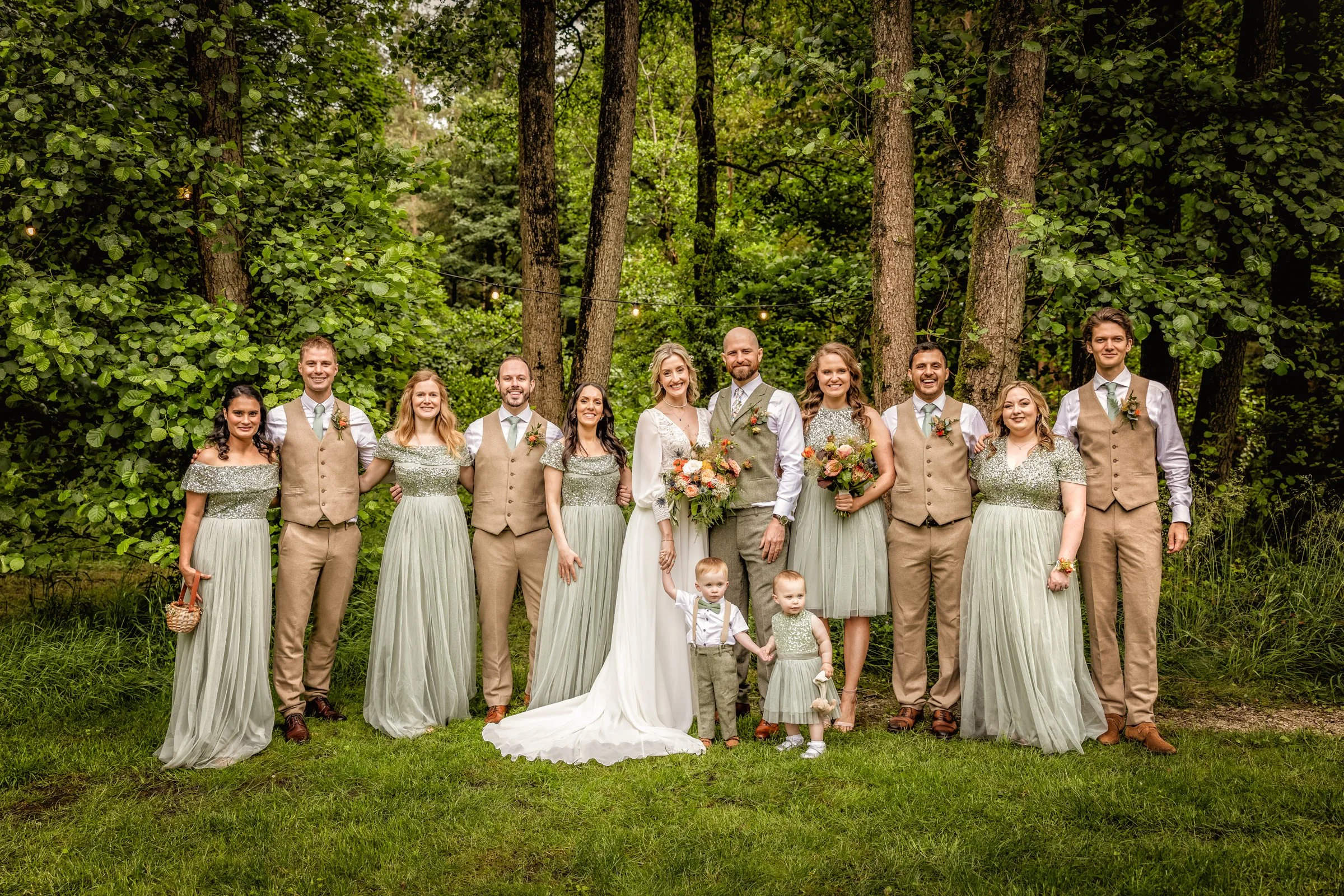 Group of 12 people, including men, women, and two children, dressed in wedding and formal attire, standing outdoors in a wooded area with green trees and grass.