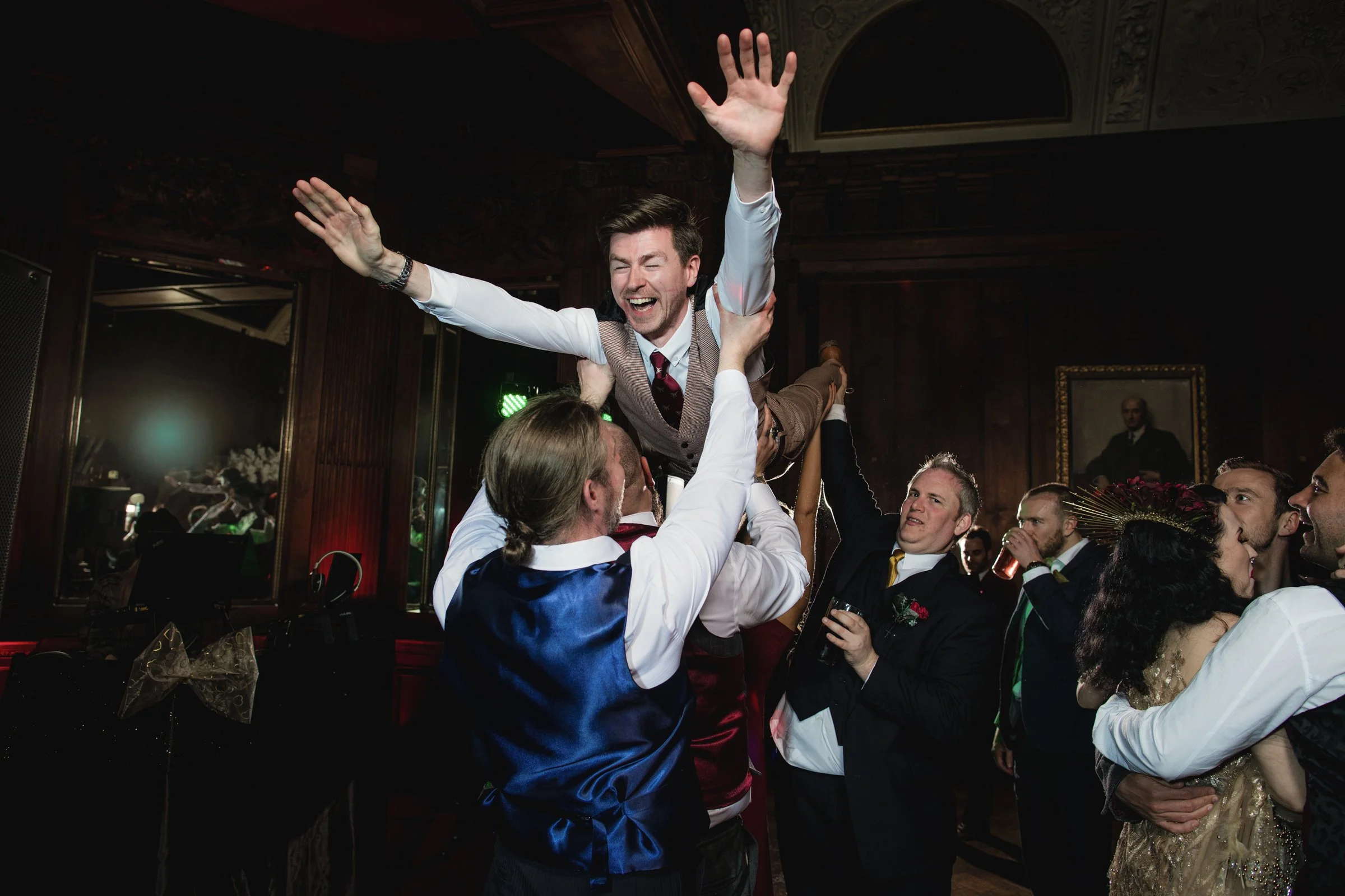 Group of people lifting a man in a celebratory setting, with some people holding drinks, in a decorated venue.