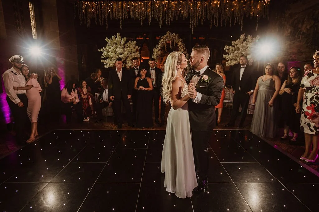 A bride and groom dance in the center of a wedding reception dance floor, surrounded by guests watching their first dance, under decorative hanging lights and floral arrangements.