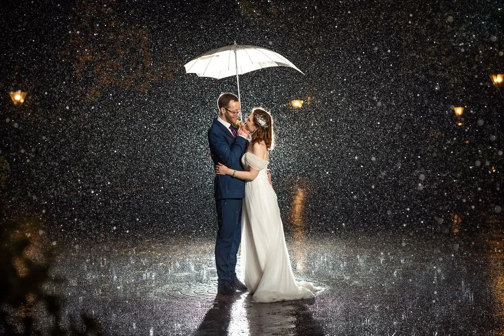 A couple stands close together under an umbrella in the rain at night, sharing a kiss. Most of the image is filled with raindrops illuminated by nearby streetlights.