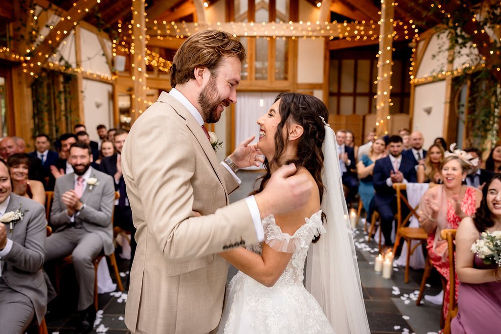Bride and groom sharing a dance at their wedding reception, surrounded by guests clapping and smiling in a decorated indoor venue with string lights.