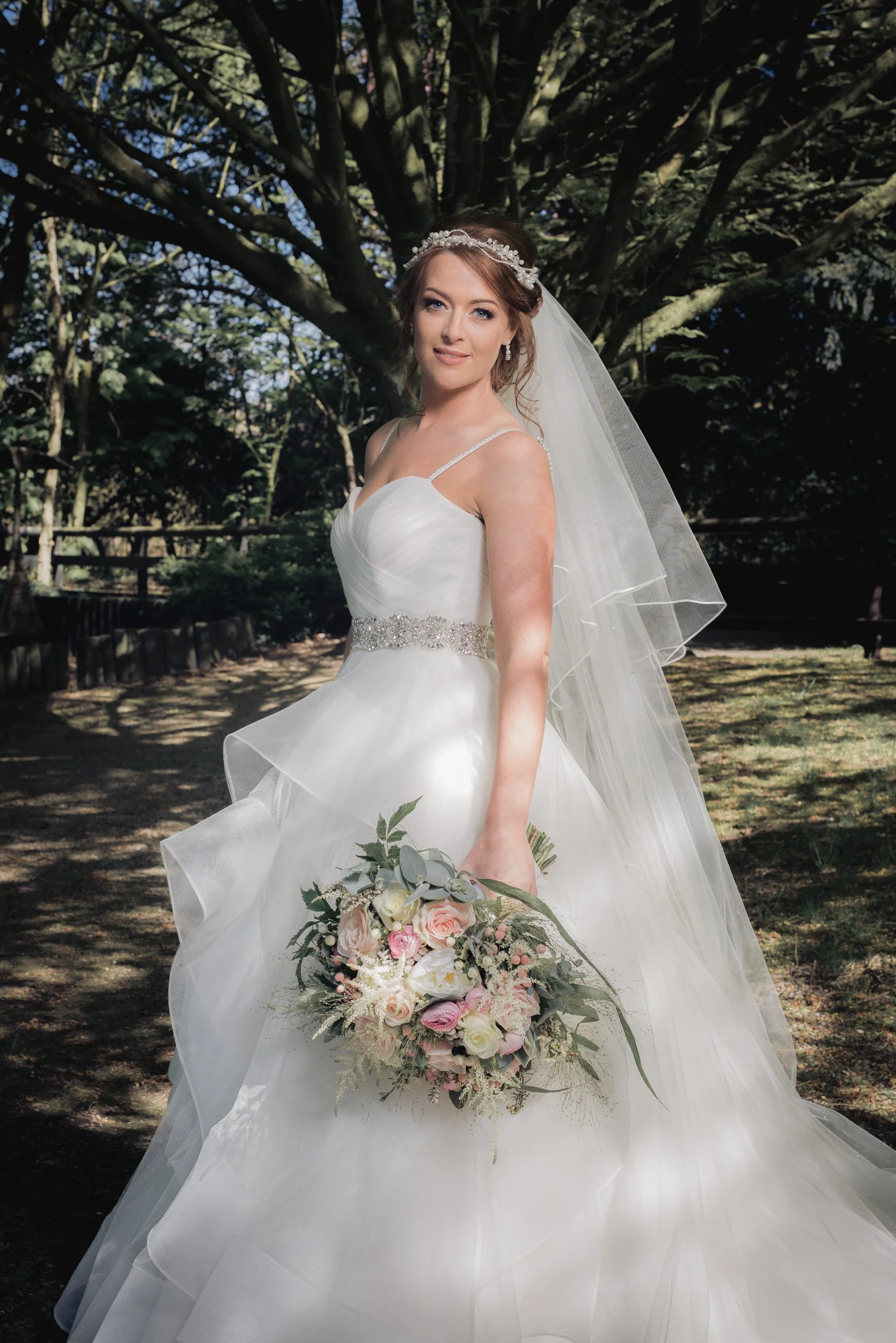 A bride in a white wedding gown with a beaded belt, holding a bouquet of pink and white roses, standing outdoors under a large tree.