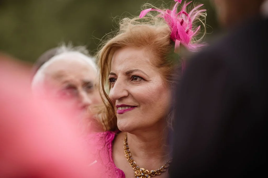 A woman with red hair, wearing a pink dress and a decorative pink headpiece, smiling outdoors surrounded by others.