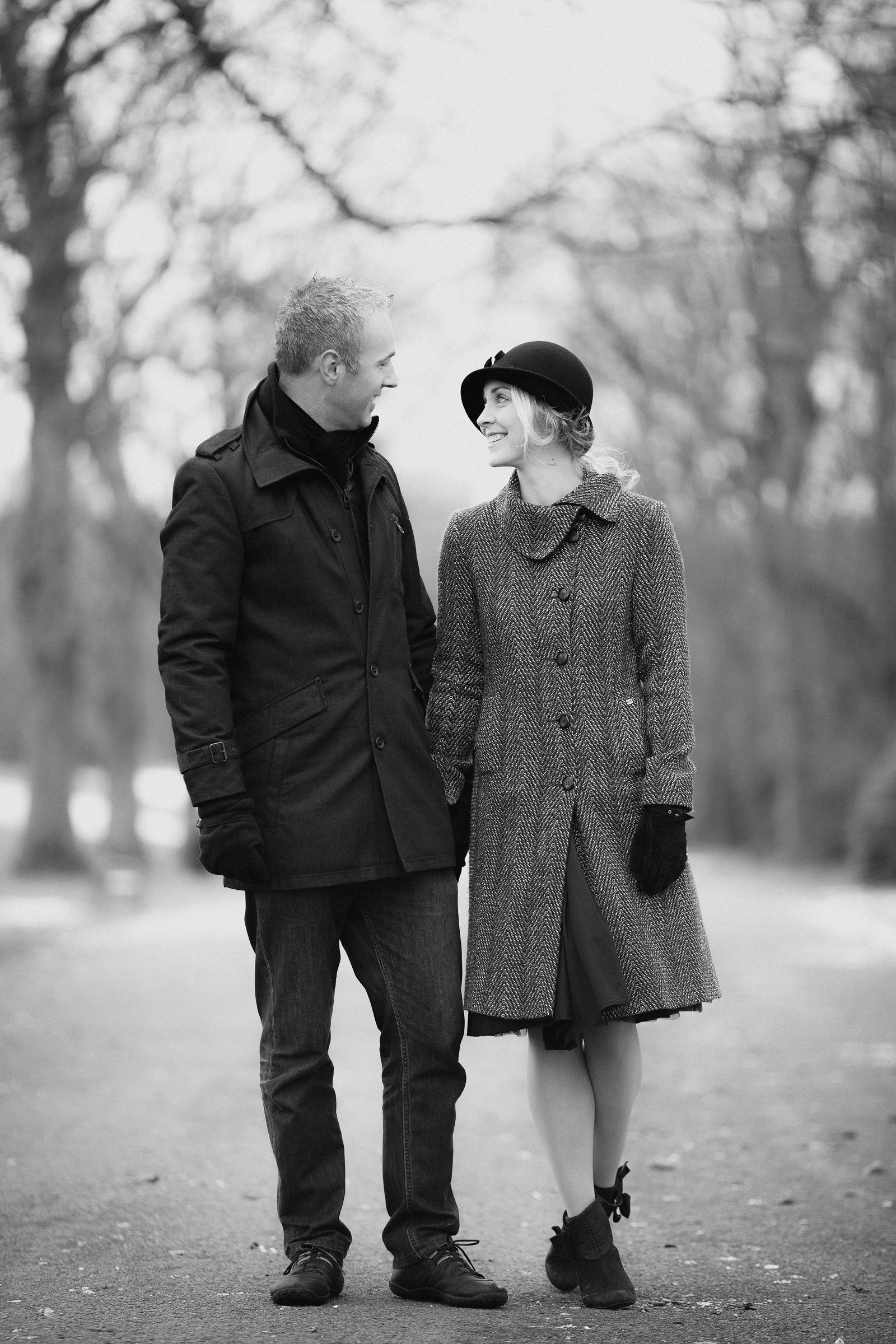 A man and woman walking together outdoors in winter, holding hands and smiling at each other, dressed in warm coats and gloves.