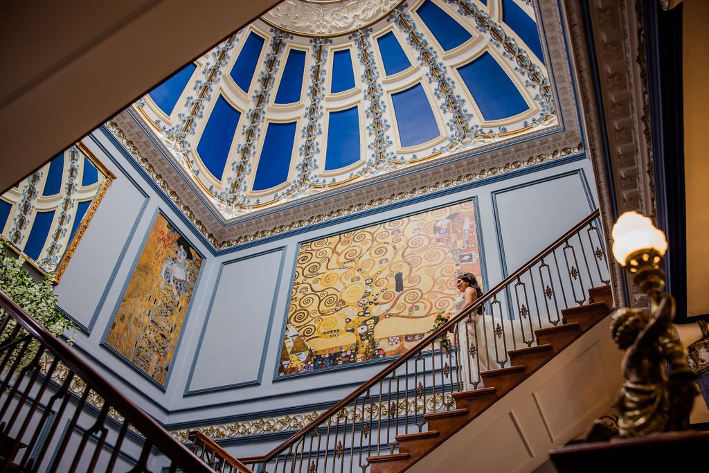 Interior view of a staircase leading to a painted ceiling and large artwork on the wall. The ceiling is decorated with blue panels and gold accents, and the artwork depicts a woman with flowers and a colorful abstract background. There are ornate rai
